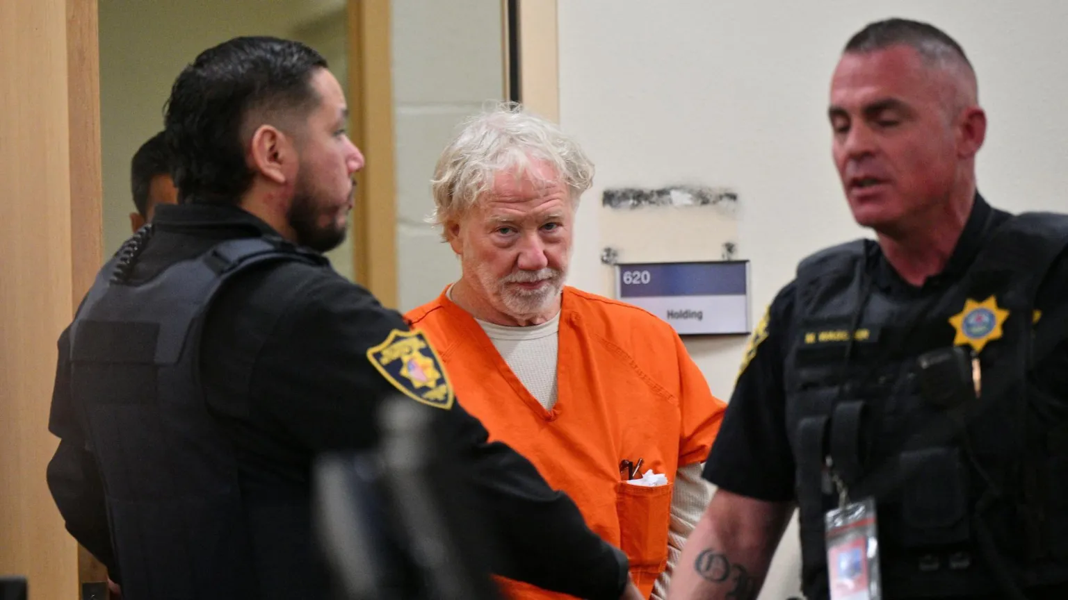  Director and actor Timothy Busfield, center, wears an orange prison uniform as he arrives for a January 2026 hearing in the Second District Judicial Court at the Bernalillo County Courthouse, in Albuquerque, New Mexico, U.S., January 20, 2026. He is flanked by two law enforcement officers in the courtroom. 