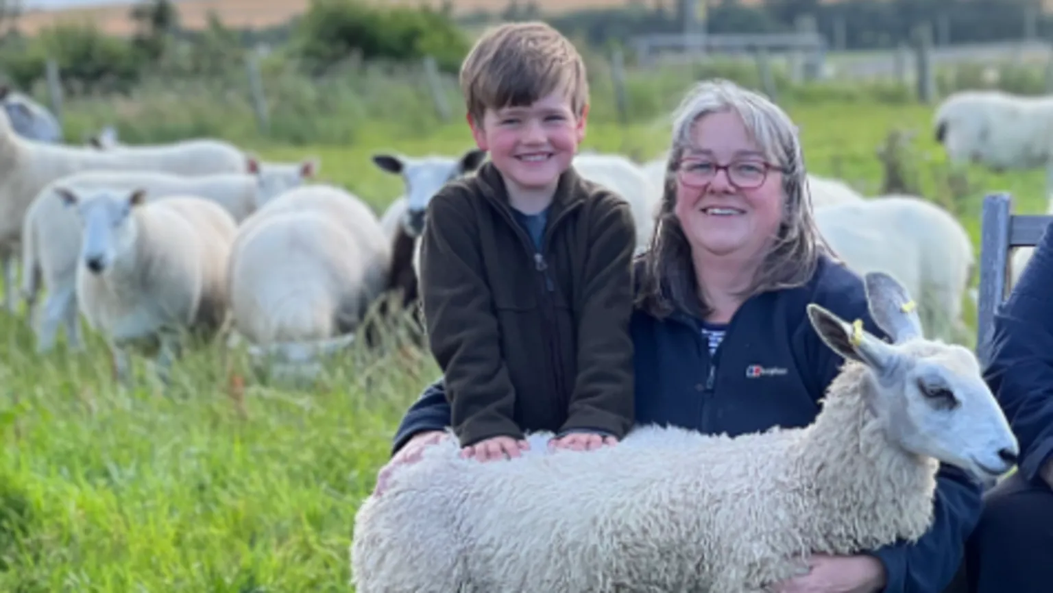 Sally Crowe Sally Crowe kneels in a field with her hand around her young son William. They are both wearing fleeces and there is a small sheep standing in front of them.