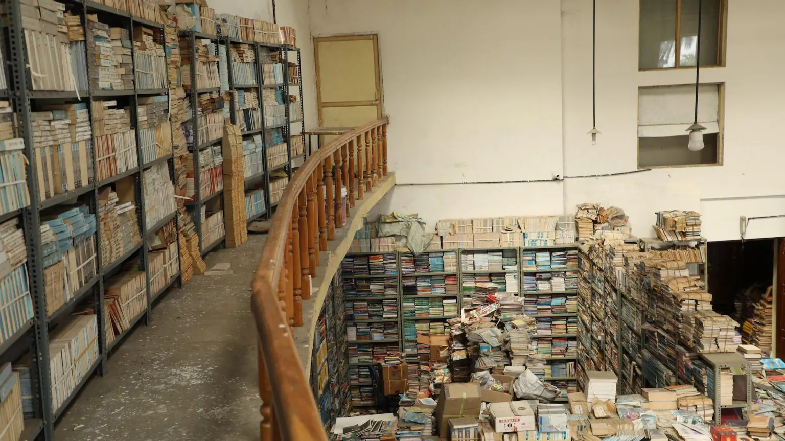 Alphonse Vimulraj The photo shows part of the upper floor of a building with books stacked neatly on shelves. On the lower floor, there are hundreds of books on the shelves and piled up on the floor.