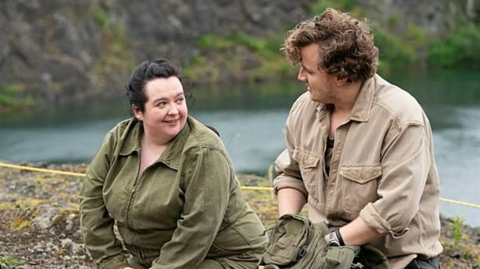 Two Brothers Productions Ashley Storrie in a scene from Dinosaur. Her character Nina is wearing a green jumpsuit and the character of Clay has a tan jacket on. They are both looking at each other while sitting on an archaeological dig site. 