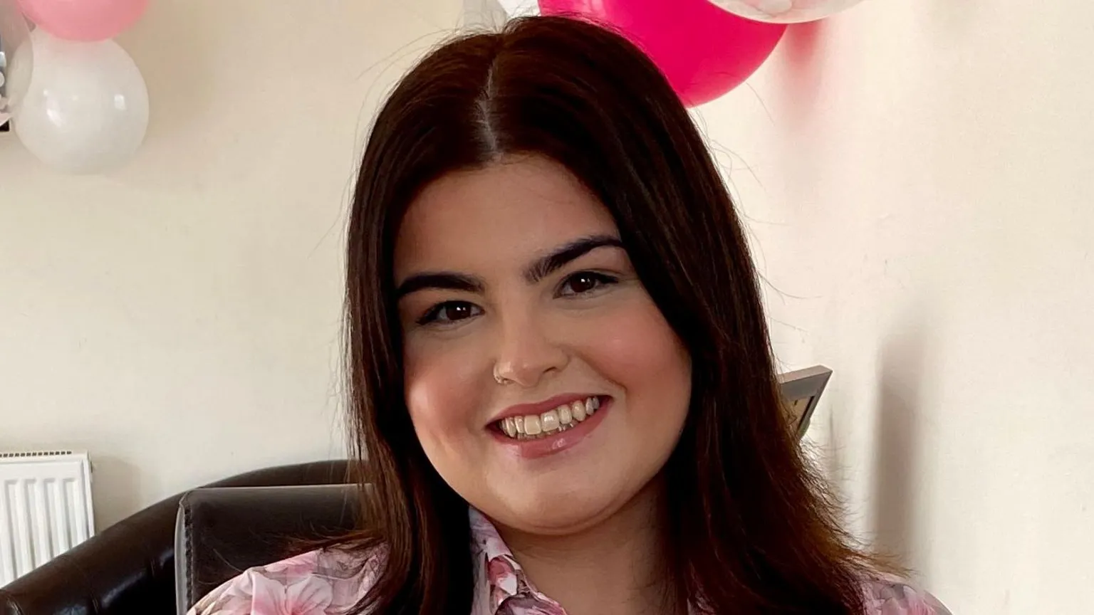 Ayla Jones Ayla Jones is a white young woman with shoulder length dark brown hair. She is wearing a floral pink blouse. Behind her on the wall are some clear, white and pink balloons and there is pink confetti on the table. She is smiling directly at the camera.