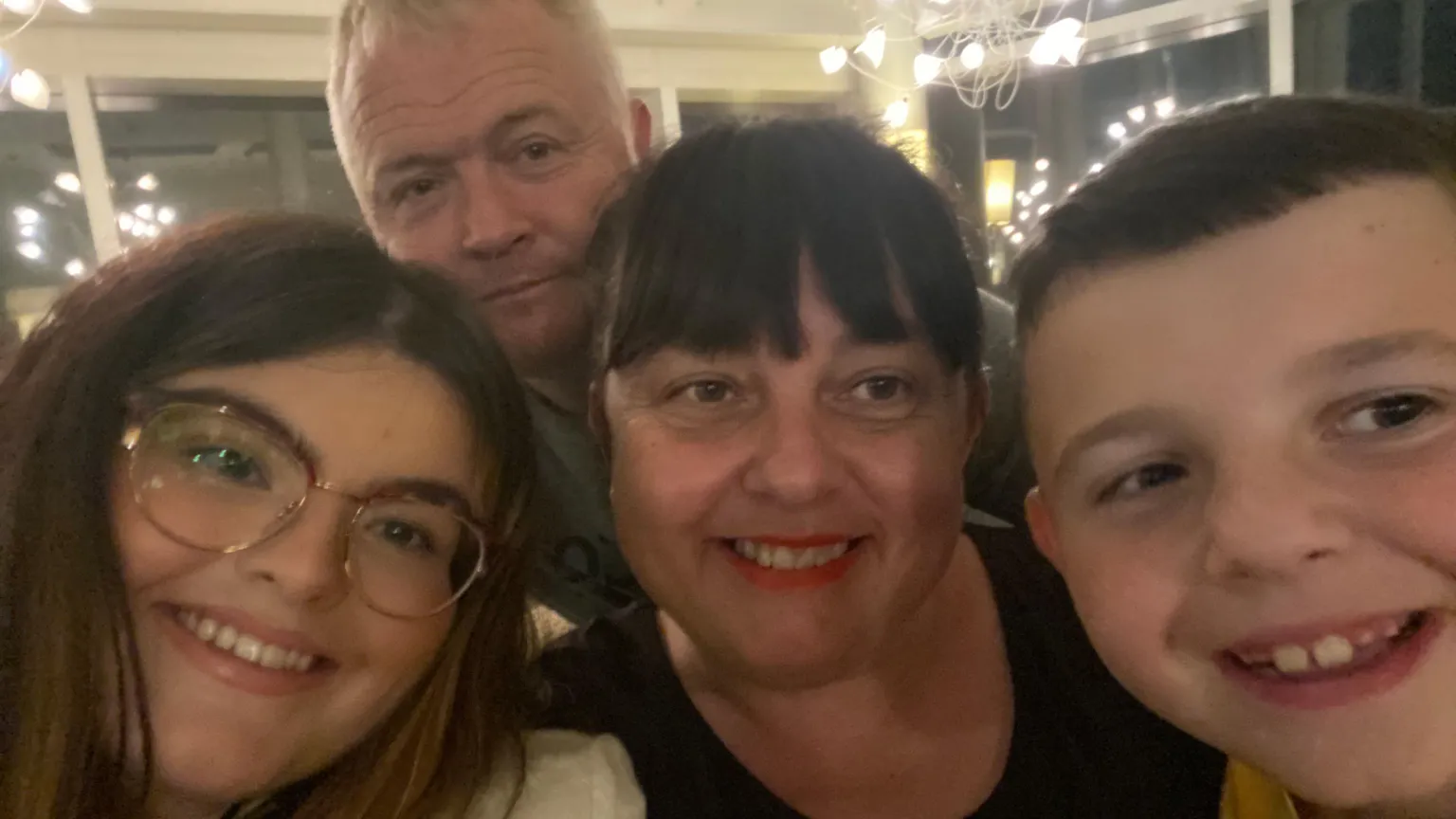 Ayla Jones Selfie photograph of Ayla (left) with her mother and father (centre) and her younger brother. The four of them all smile and look directly into the camera lens. Alya has brown hair, brown eyes and wears reading glasses. Her mother has a dark brown full fringe and brown eyes, while her father stands behind them in the background and smiles. He has silver short hair.