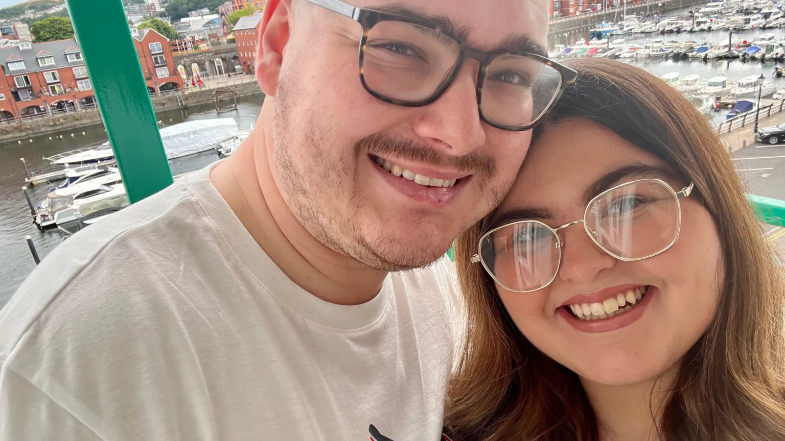 Ayla Jones White man in his 20s with a white t-shirt and dark tortoiseshell framed glasses leans his head onto Ayla Jones's. She has long brown hair and wears gold-framed glasses. It is a head and shoulders shot of them both smiling at the camera in a selfie pose. They appear to be high up with windows behind them and in the background the viewer can see Swansea Marina underneath with rows of converted warehouse flats and multiple small boats and yachts moored in the water.