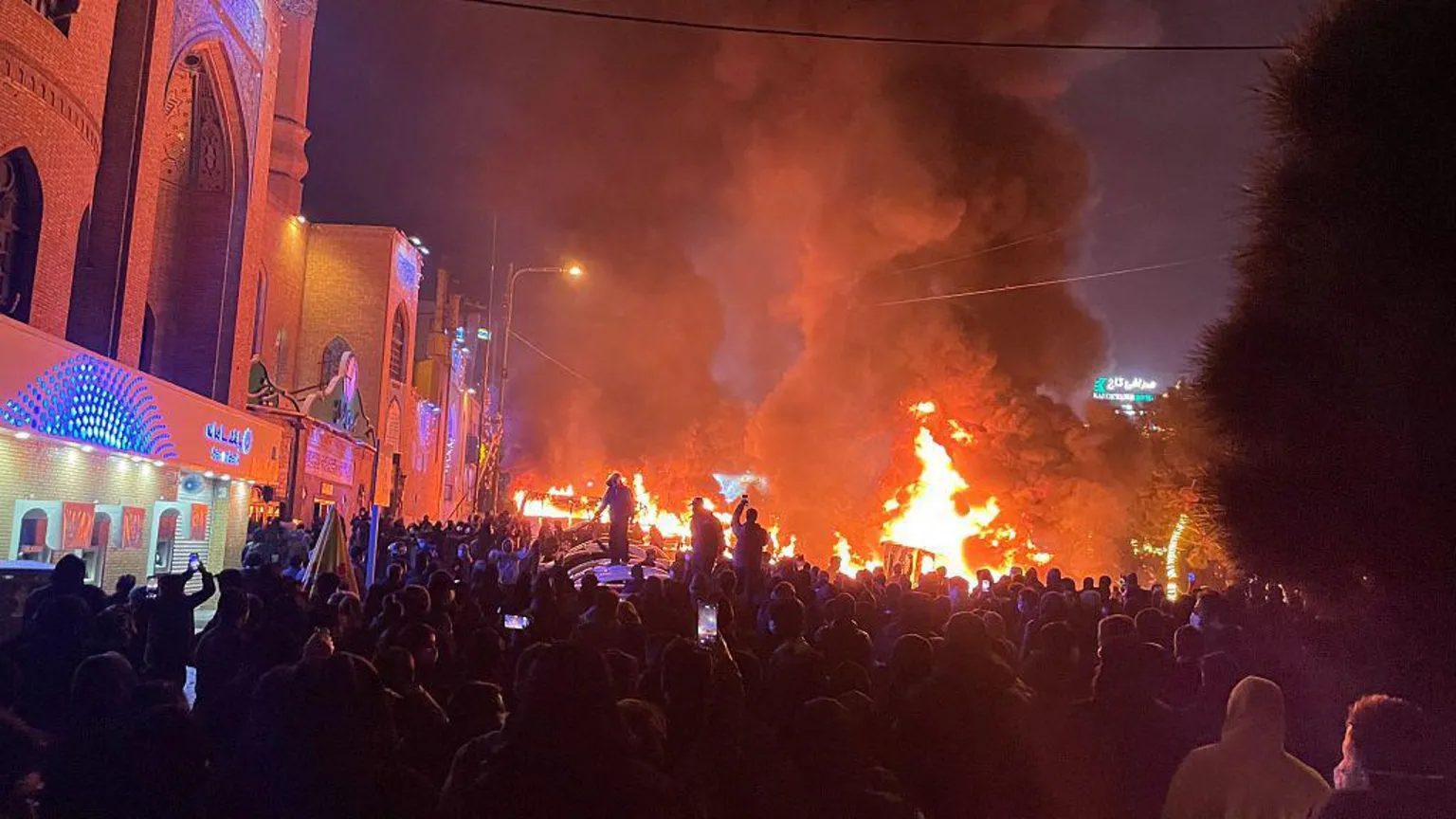 AFP via image of dozens of protesters gathered on a street in Tehran, Iran as cars burn in the background