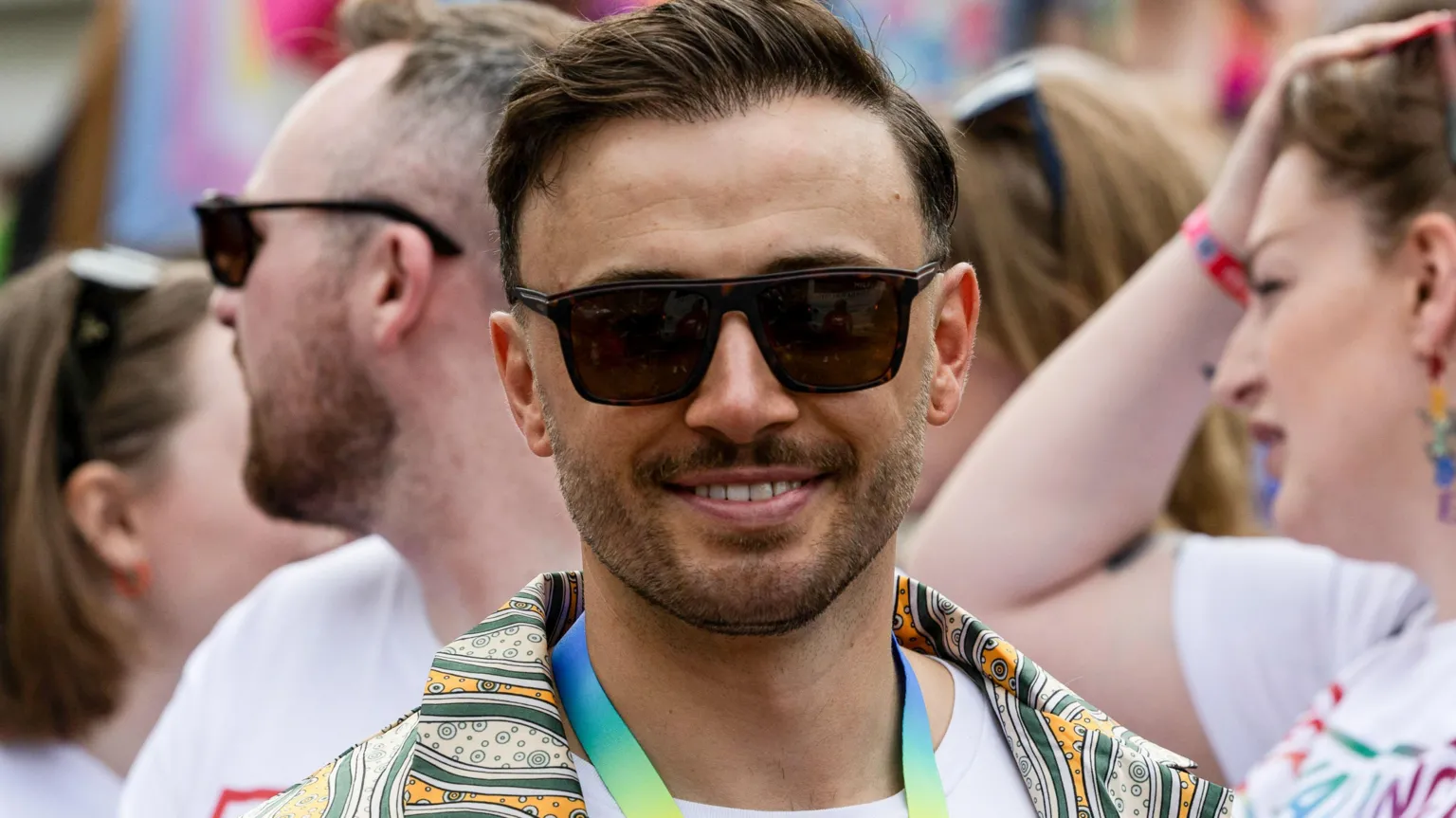 SNS Zander Murray stands in a crowd at Edinburgh Pride 2023. He has dark hair and a beard and is wearing sunglasses and a rainbow lanyard around his neck. He is also wearing a green and yellow patterned shirt.