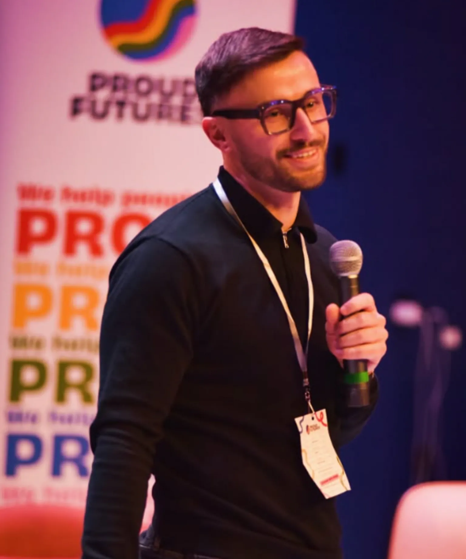 Zander Murray Zander Murray, who has dark hair and a beard, is standing in front of a huge Pride sign. He is wearing a black, long-sleeved shirt, a lanyard around his neck and glasses. He is holding a microphone and smiling.