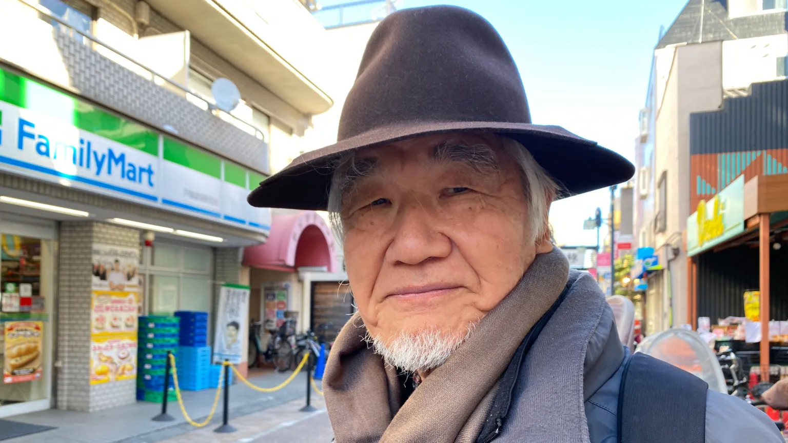 Naoaki Yuhara, in a wide brimmed hat and scarf, stands in front of a FamilyMart in Tokyo
