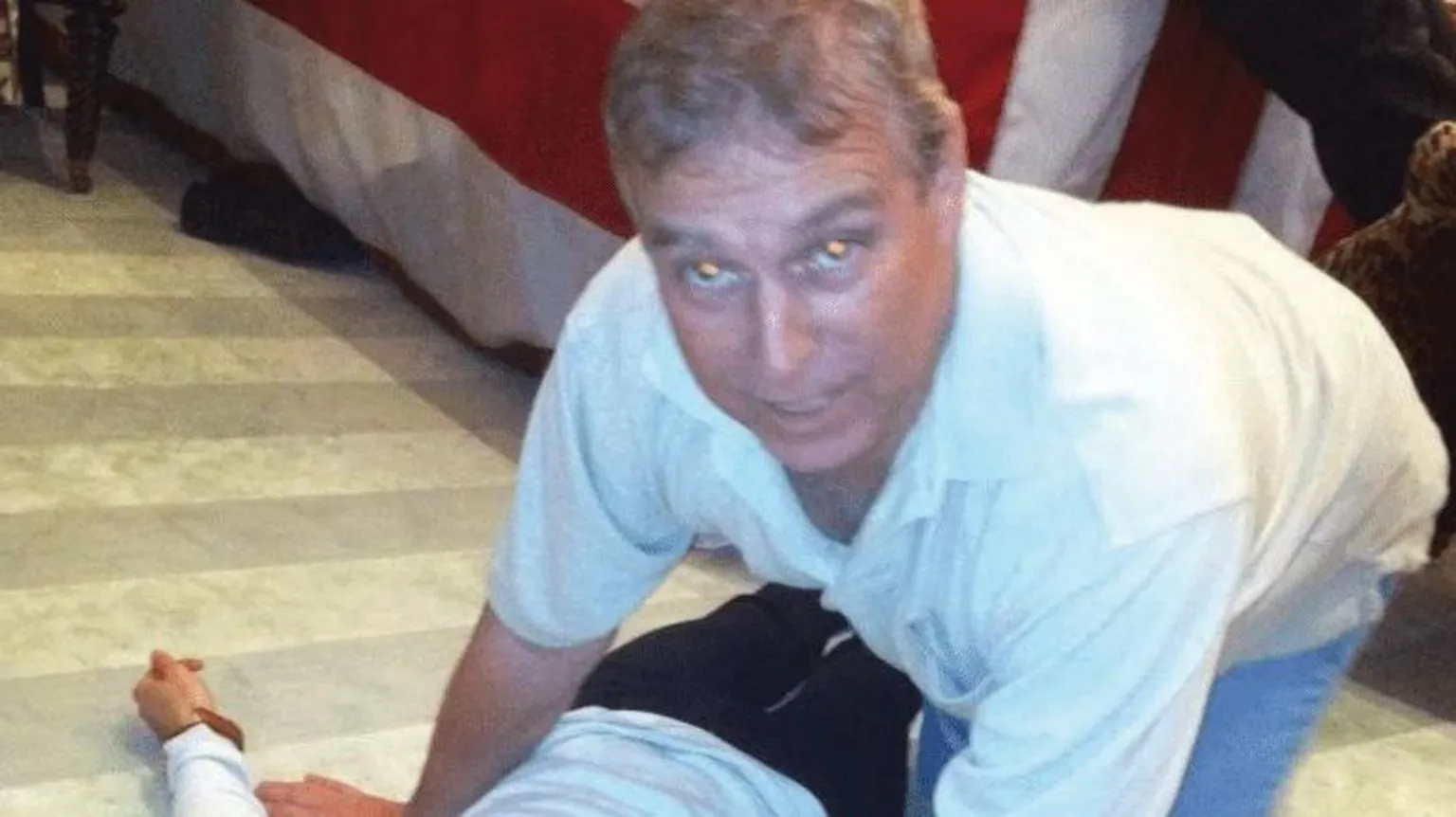 US Department of Justice A grey-haired man dressed in a white polo shirt and blue jeans looks directly at the camera. He is on all fours directly over an unidentified female who is laying on the floor.
