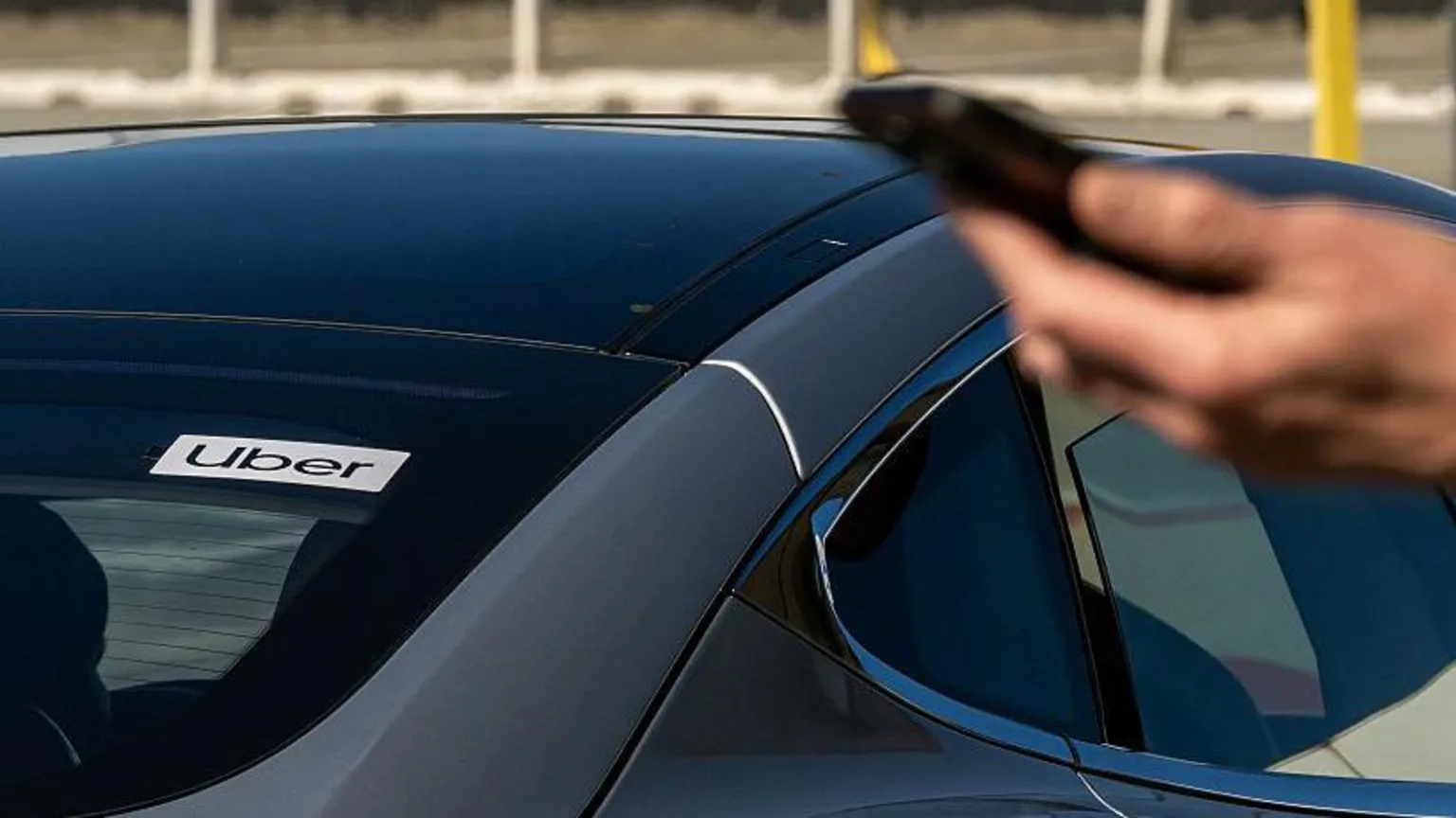 Bloomberg via A male hand holding a mobile phone in the foreground with the back of a black car behind him. In the rear window of the car there is an Uber sign.
