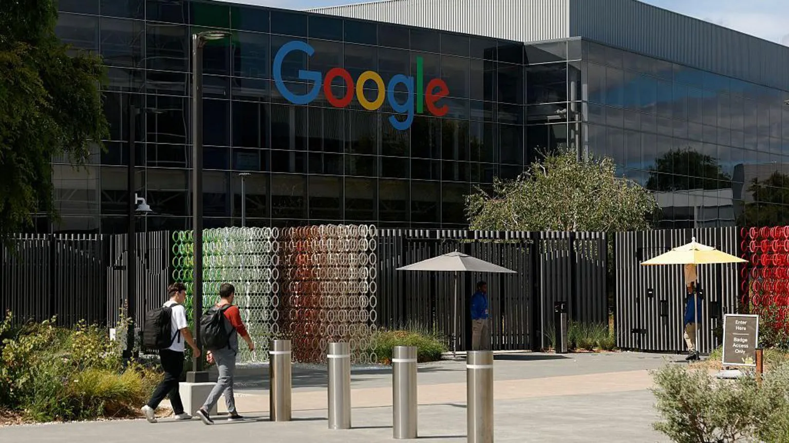  Outside of a large dark-coloured shiny building with the Google logo in blue, red, yellow and green letters high up on the wall. In front of it two young unidentifiable men with large backpacks are walking with their backs to us