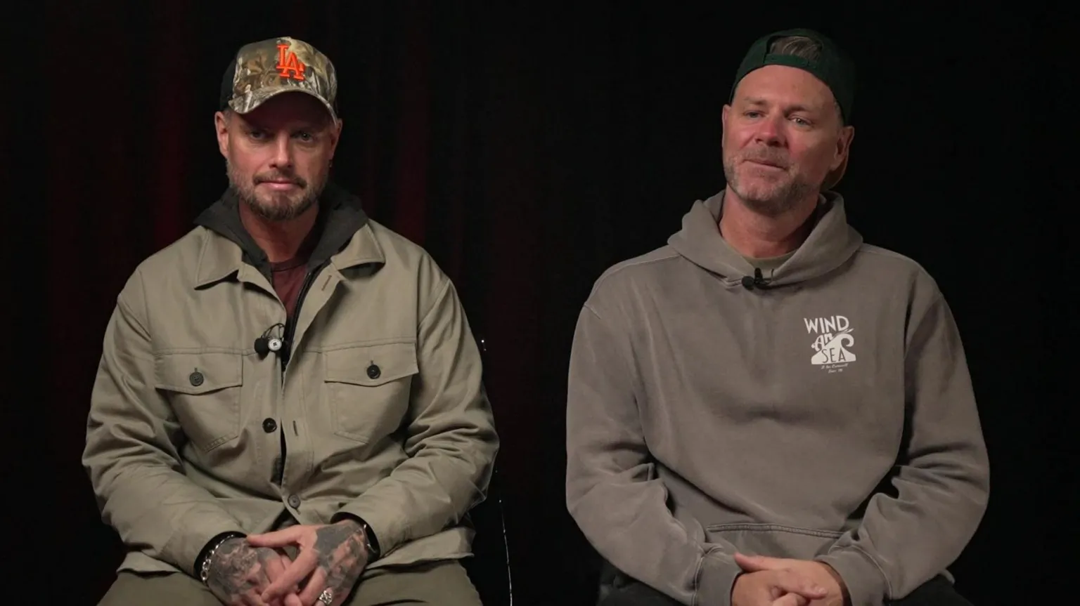 Two men sit side by side against a dark background. Both are casually dressed in hoodies and jackets, wearing baseball caps. They face the camera, hands resting in their laps, appearing to be in an interview or studio setting.