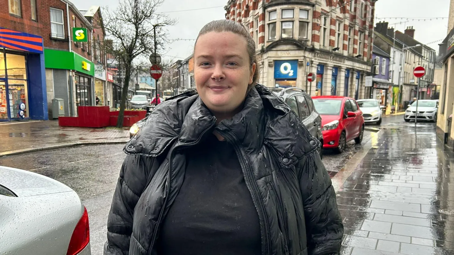 BBC A woman stands close to the camera on a very rainy day. She has brown hair tied back in a ponytail. She is wearing a black puffer coat. The pavement is slick and reflective in the rain behind the woman, backed also by a line of traffic. Behind her is a busy street in strabane, with buildings like a subway and o2 shop in vision. 