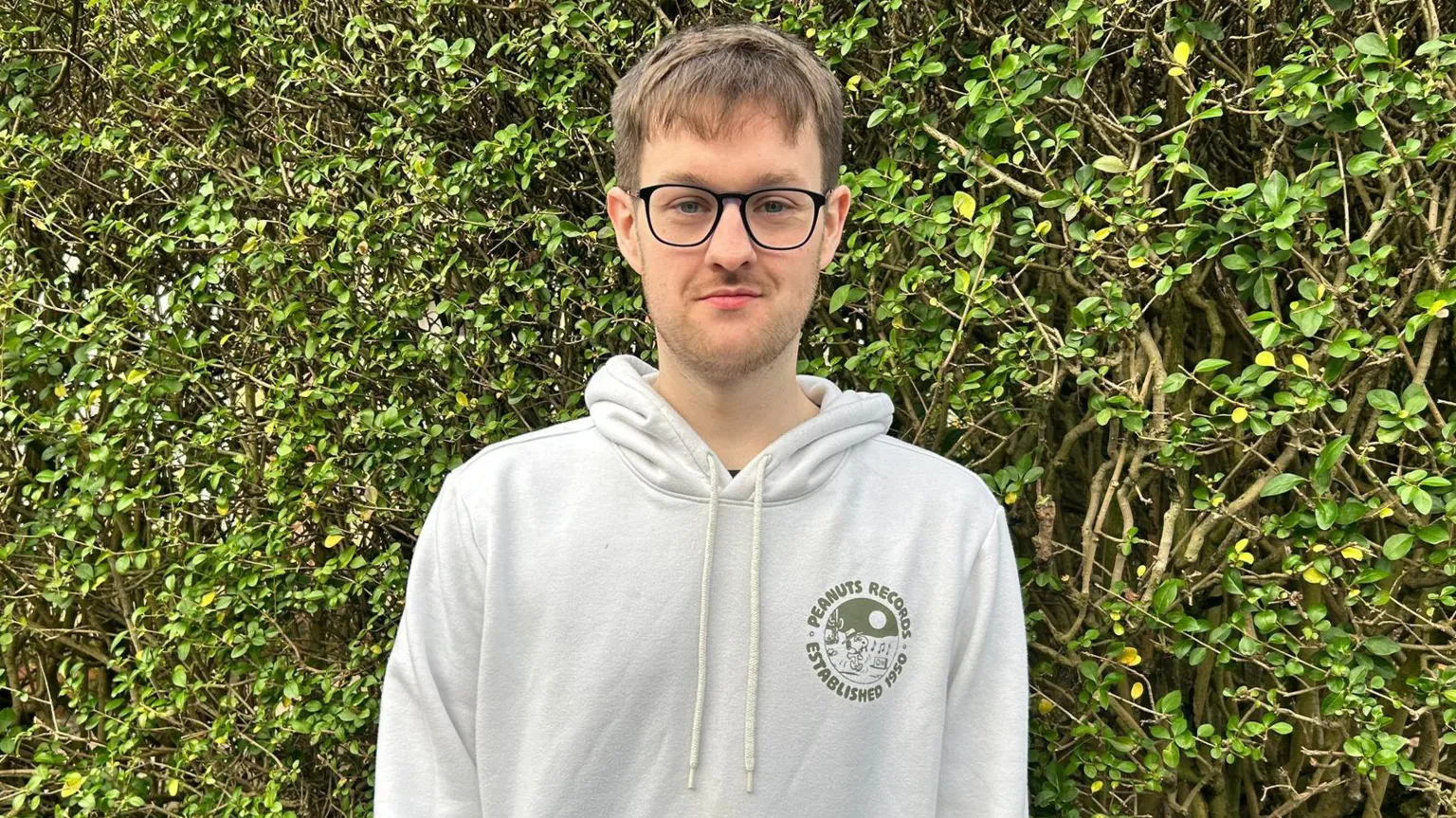 A man stands close infront of a green hedge. He has short brown hair, with the beginnings of a 5 o'clock shadow. He is wearing black glasses with large lenses, and a white hoodie with a round graphic on the left of his chest.