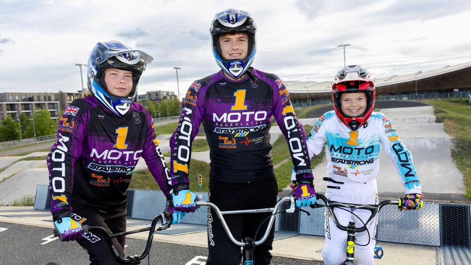 Better.org.uk The three siblings stand with their bikes with the BMX track behind them. Katie and Thomas wear purple racing suits with a blue helmet and Phoebe wears a white and blue suit with a red helmet