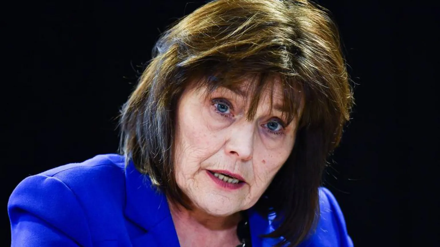 Jeane Freeman, a woman in her 60s with shoulder length dark brown hair, is wearing an electric blue jacket and is sitting talking against a black background.