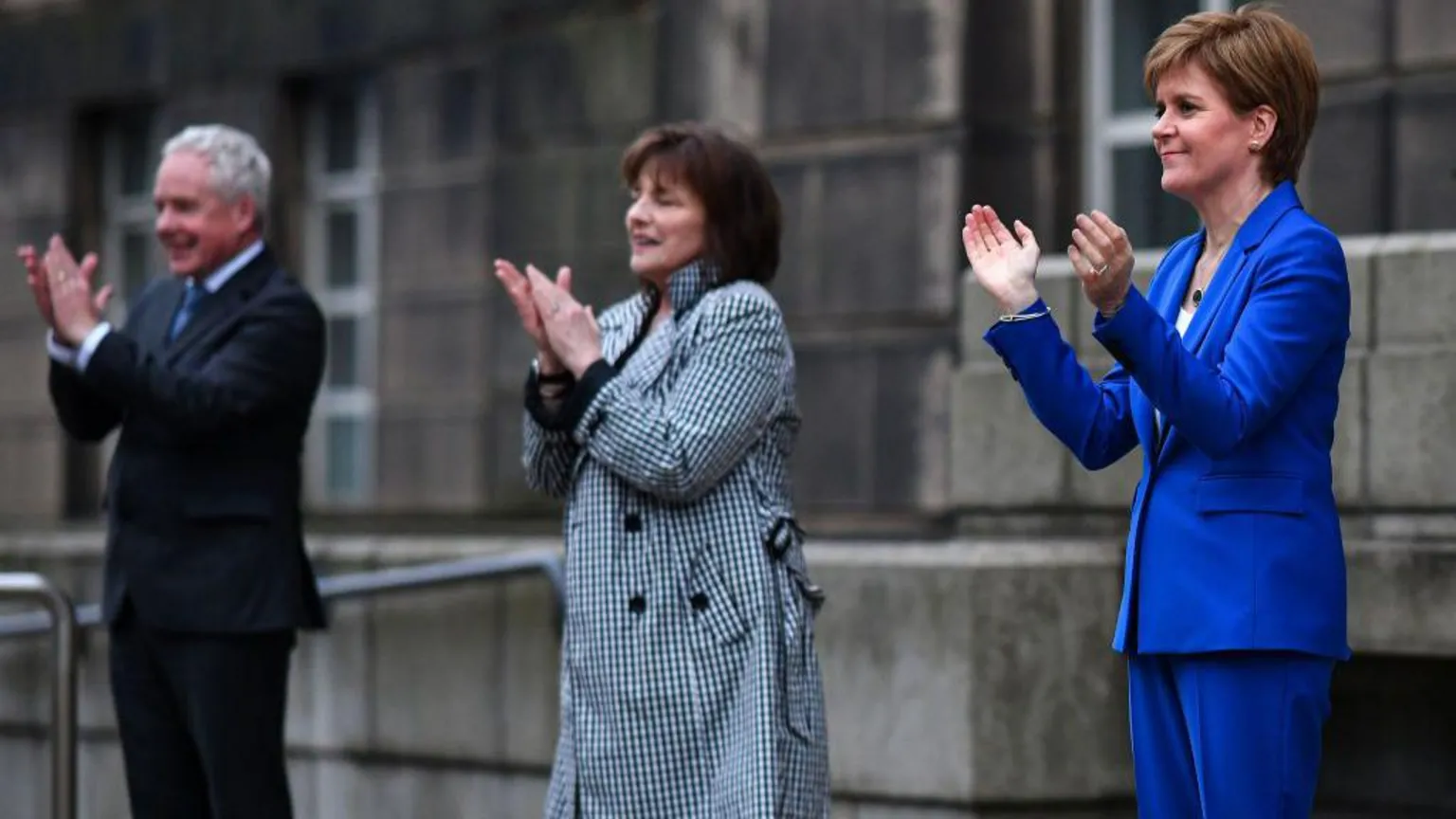Malcolm Wright, a man in a dark suit; Jeane Freeman, a woman in a black and white checked coat and Nicola Sturgeon, a woman in an electric blue trouser suit, stand in a row applauding outside a grey stone building (St Andrew's House).
