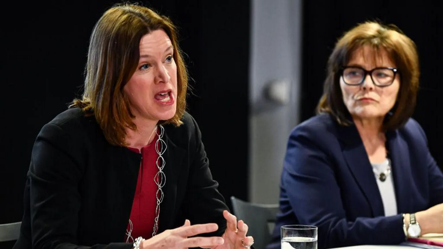 Catherine Calderwood, in black jacket over a red shirt, is sitting at a desk talking. Jeane Freeman, in dark blue jacket with a white shirt, is sitting to her right watching her speak. Freeman is wearing spectacles with thick black plastic frames.