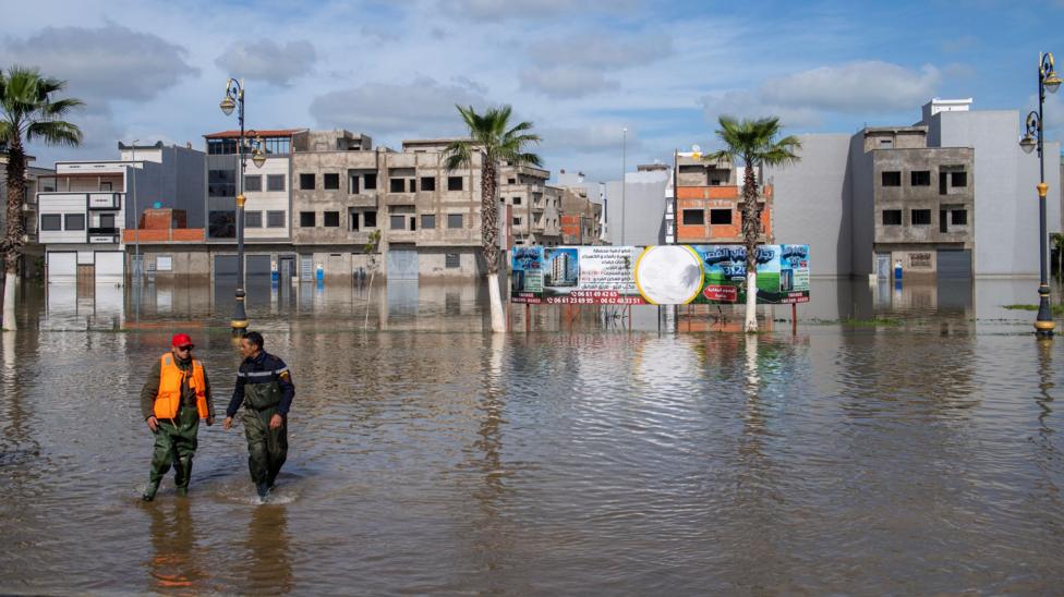 Buildings and palm trees. Two men wade in a flooded street.
