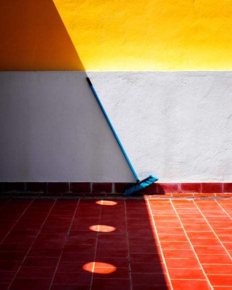 A broom leaning at an angle against a wall, its stick aligning with the shadow on a yellow wall above, with red tiles below