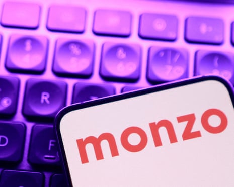 Red Monzo logo on a white phone screen seen against a backdrop of a computer keyboard with black keys, illuminated in purple. 