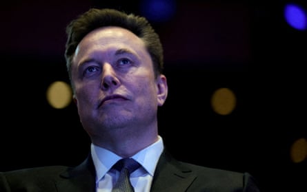Elon Musk in head and shoulders image against a dark background: he wears a dark jacket and tie with white shirt and is holding his chin up, looking serious.