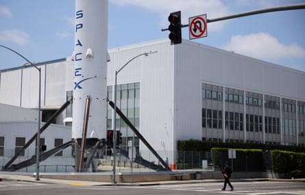 View of the corner of a white, industrial-looking rectangular building with a Space X rocket at the front by the entrance, seen from a road crossing with overhead traffic lights. 