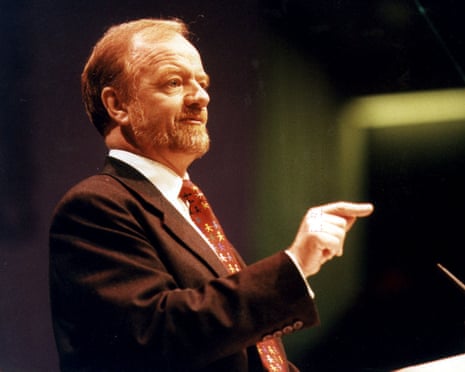 Robin Cook addressing the 1997 Labour party conference in Brighton: he wears a brown jacket and red tie, and points a finger. He has short reddish hair and beard.