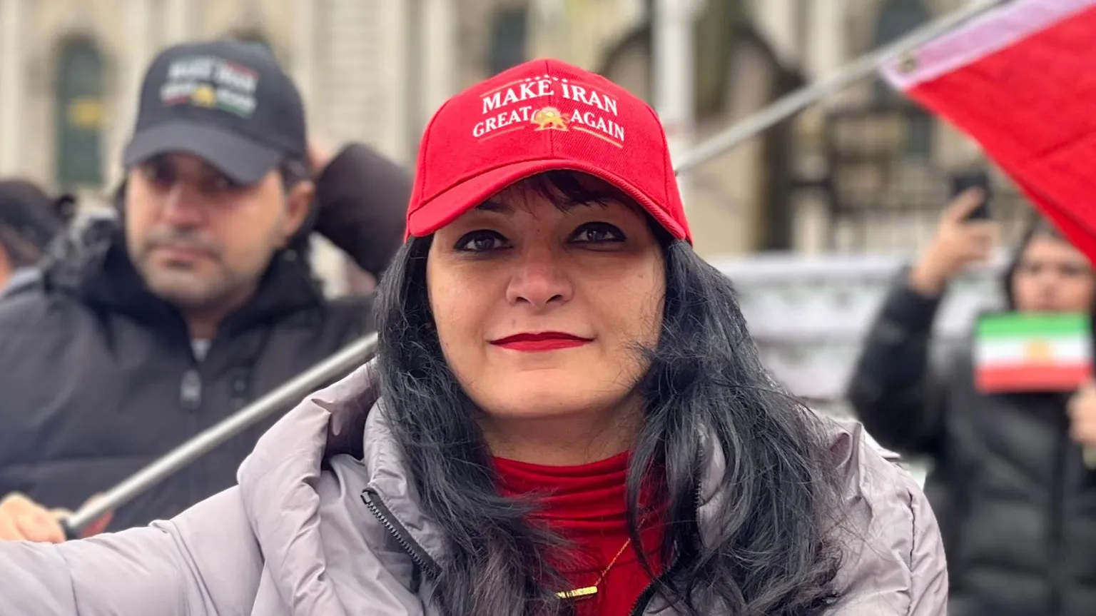 BBC A woman with black hair is standing outside at a protest in front of a building. She is wearing a red hat, top and grey coat. Other people can be seen wearing coats behind her. A small Iranian flag can be seen held by someone out of focus behind her.