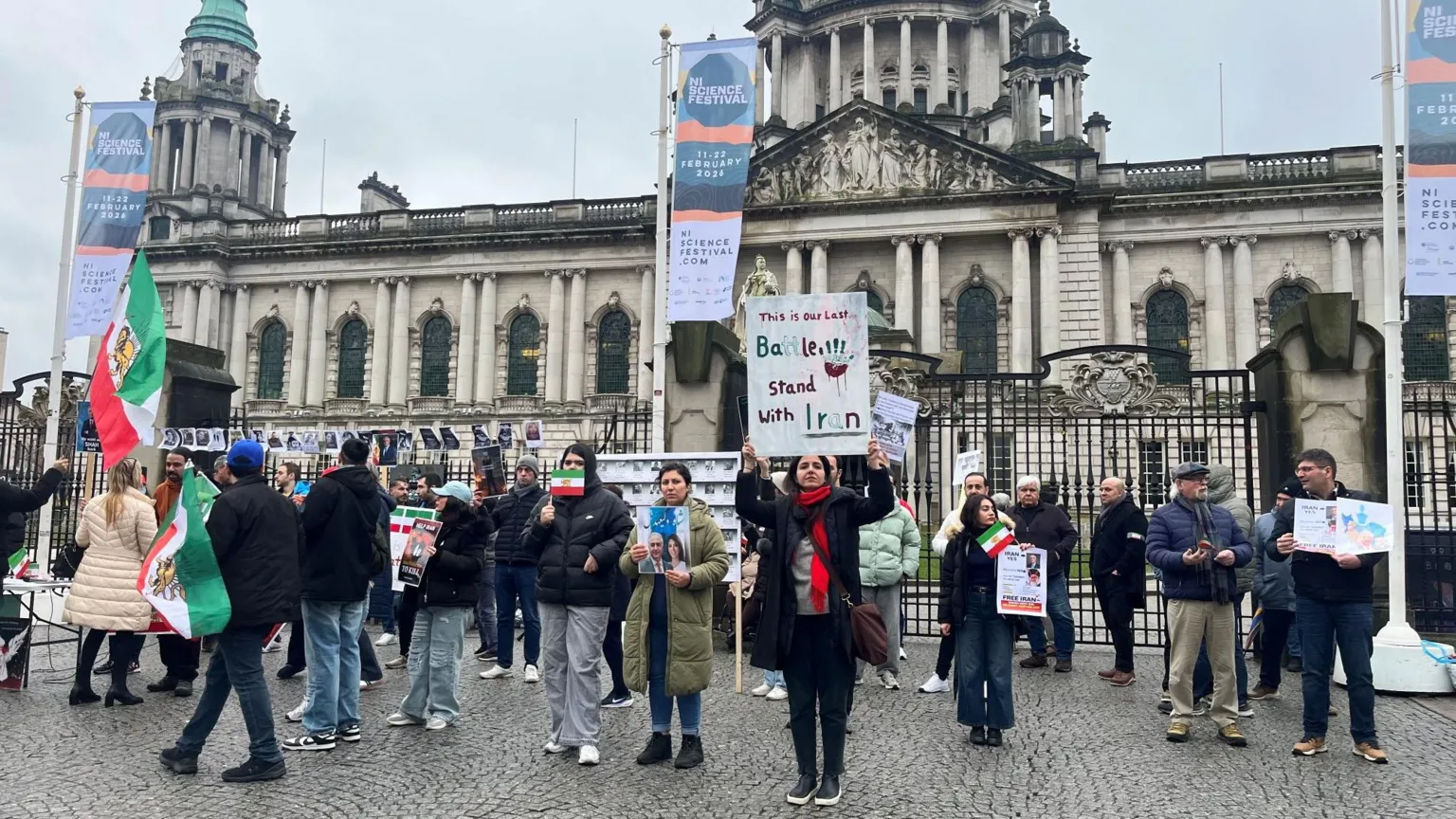 Protesters stand outside in winter attire in front of a big grey ornate building. They are holding various banners and Iranian flags.