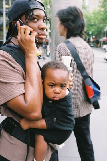 a young man talks on a phone whilst holding a baby on his chest in a baby carrier 