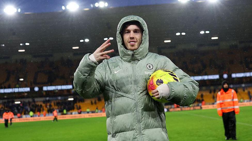 Cole Palmer celebrates scoring a hat-trick for Chelsea with a three-finger salute with his right hand, while holding the yellow match ball under his left arm and wearing a large Chelsea puffer jacket with the hood up.