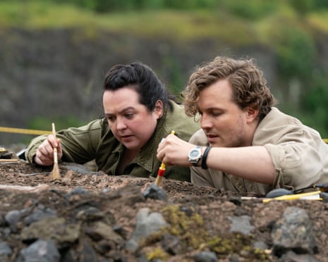 Ashley Storrie and Hyoie O’Grady standing inside an earth hole, using small brushes to uncover something