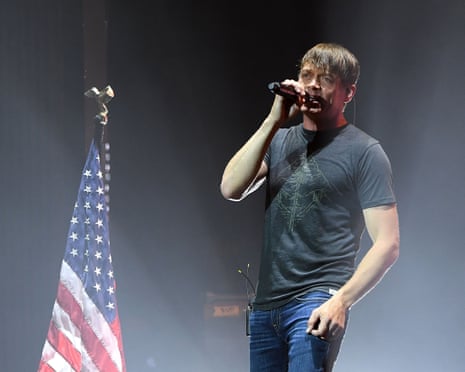 Man on stage in T-shirt and jeans in front of US flag.