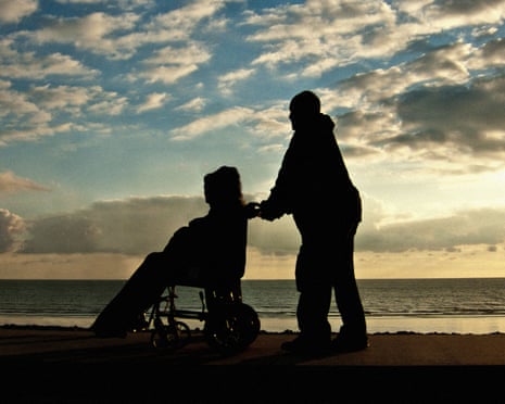 A silhouetted figure pushes another in a wheelchair on a seafront