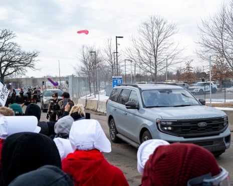 A pink dildo and a purple dildo sail through the air at a greay SUV driving alongside a crowd of protesters.