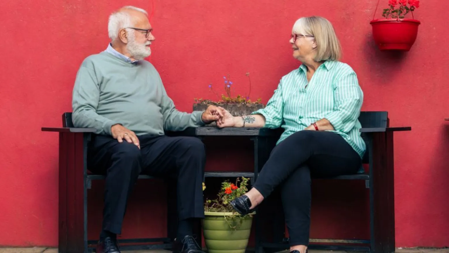 Curious North Productions An older couple sits on a wooden bench against a bright red wall, holding hands across a green planter filled with flowers. The man has grey hair and a full beard, and wears a light green sweater and dark trousers, while the other woman wears a green‑striped shirt and black trousers. A red hanging pot with flowers is attached to the wall above them. 