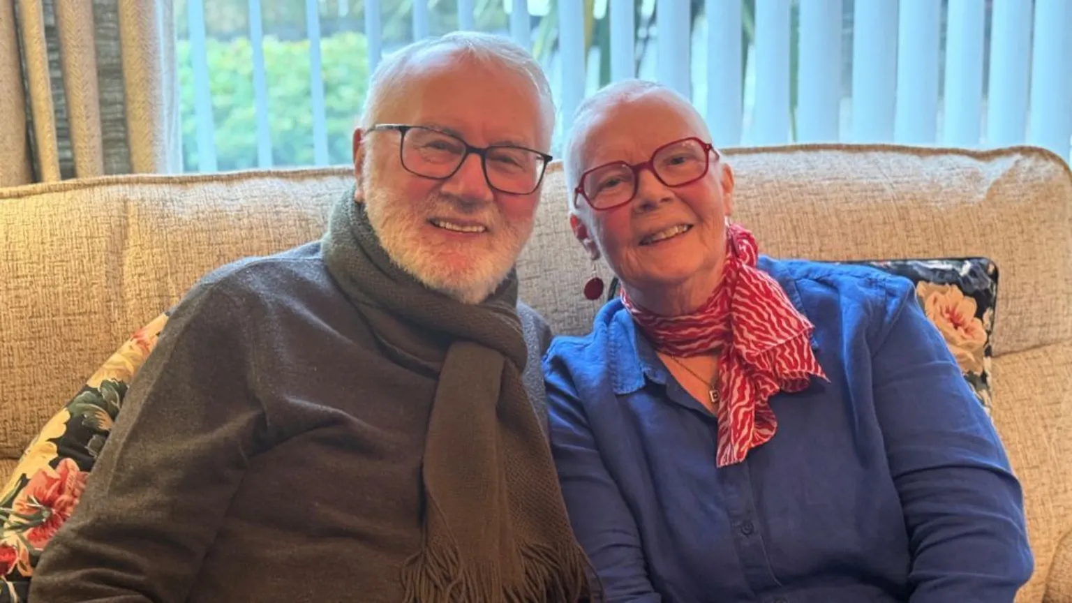 Two people sit close together on a beige sofa in a warmly lit living room. The man with grey hair and beard wears a dark sweater and a thick brown scarf, while the woman wears a bright blue top with a red patterned scarf. Vertical blinds and greenery outside a window are visible in the background, and floral cushions rest on either side of them. 