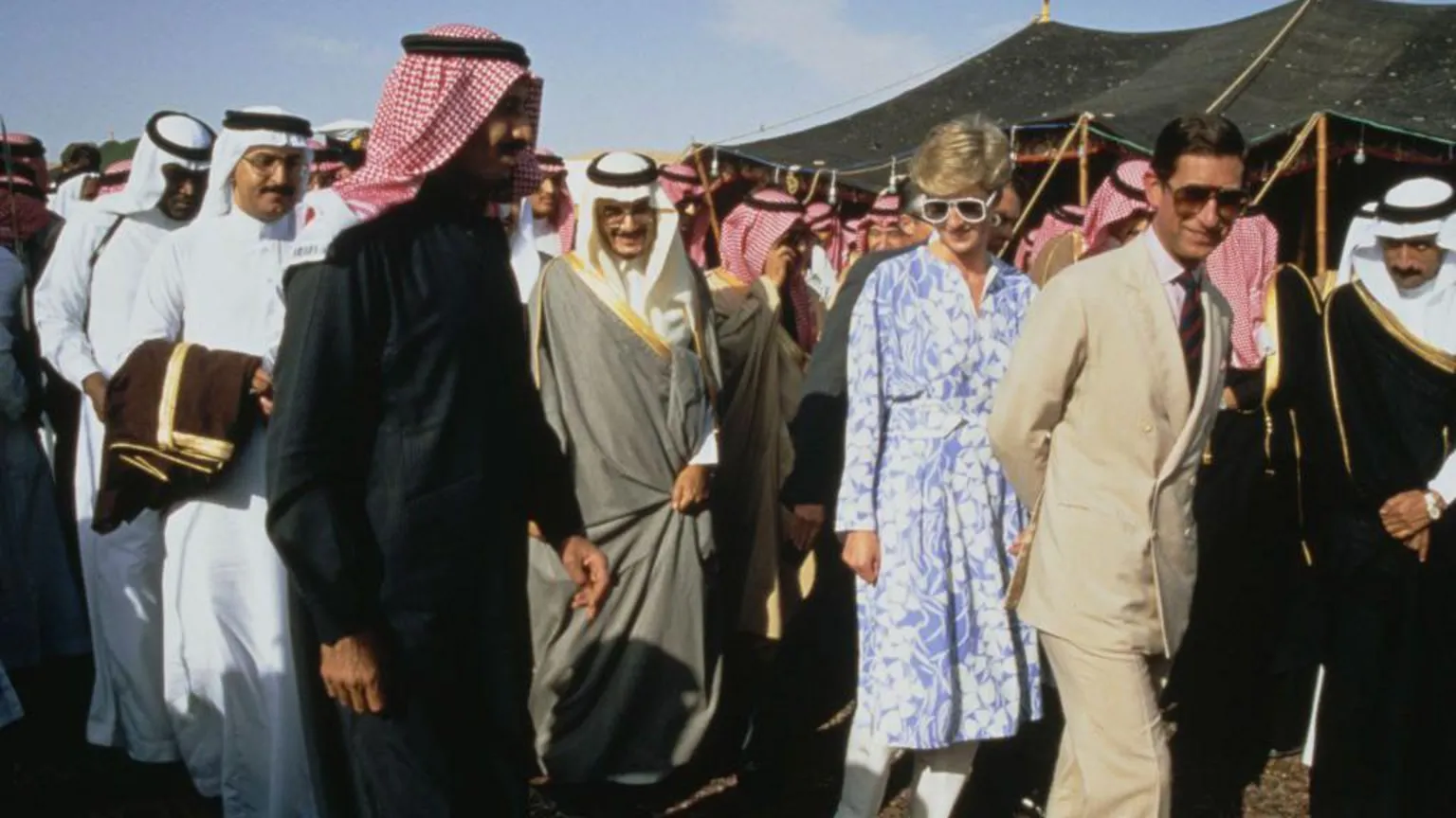 Tim Graham Photo Library via Charles, wearing a cream linen suit and sunglasses, and Princess Diana, wearing a blue and white floral dress and sunglasses, walk along smiling, surrounded by many men in traditional Saudi attire, also smiling.