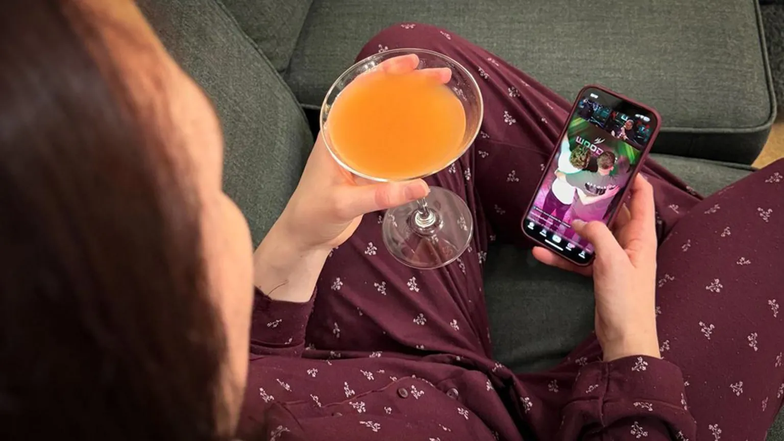 BBC Woman sat on a sofa holding a cocktail in one hand and her phone in the other. She is watching a live stream from a bar in Manchester. (Stream image has been added to phone by Photoshop for illustration purposes)