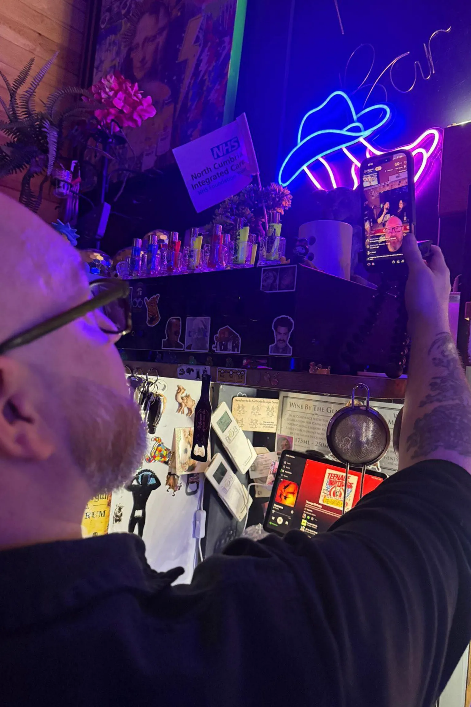Morgan's Arcade Bar Morgan attending to a smartphone recording the livestream in his bar. The phone is held in a tripod and situated on a shelf next to bottles of vodka. A bright neon sign shaped like a cowboy hat glows blue and pink above the shelf.