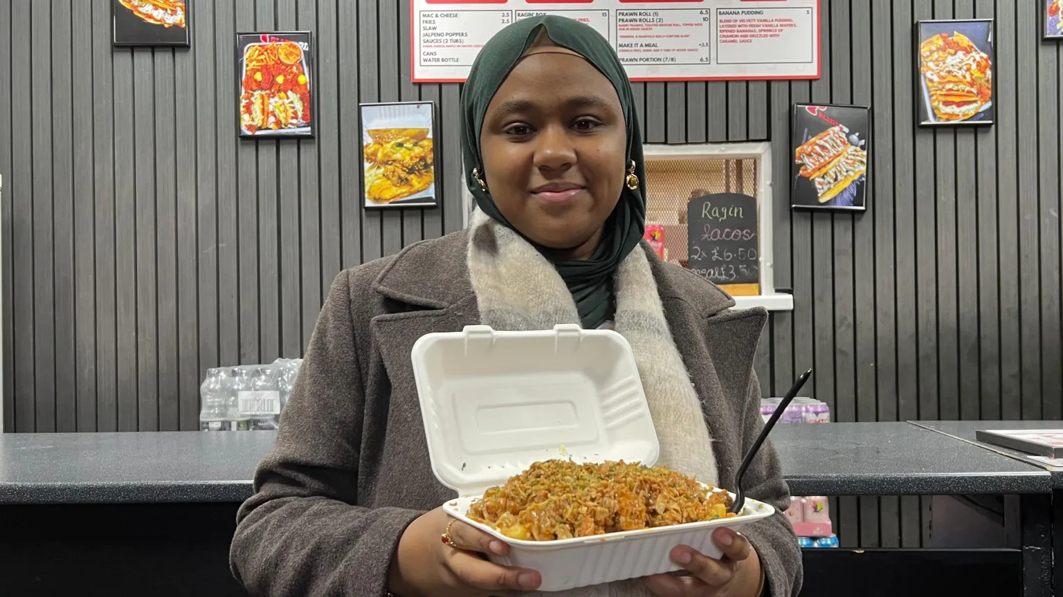 Sumayyah is wearing a beige-grey long coat and a green hijab or headscarf. She is holding an open white takeaway box filled with seasoned fried chicken inside a fast‑food shop, with menu boards and food photos displayed on the dark-paneled wall behind the counter.