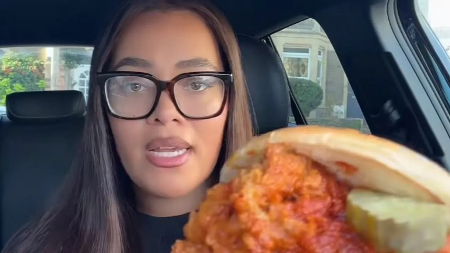 @til.eats Till has long brown straight hair and is wearing oversized specs on her face. She is sitting inside a car holding up a fried chicken sandwich close to the camera, showing a crispy chicken fillet, pickles and a toasted bun, with a residential street visible through the car window in the background.