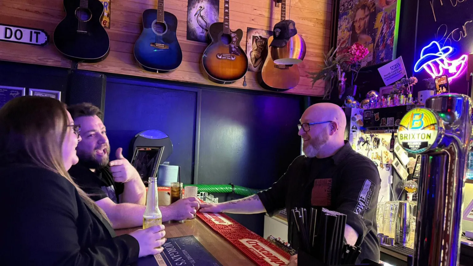 Morgan's Arcade Bar Two people stand at Morgan's bar counter holding drinks while speaking with him. The bar surface has branded mats, straws, and drink taps visible. Behind the bar, shelves contain bottles, decorations, and a neon sign shaped like a hat. Several guitars hang on the wooden wall above the seating area.
