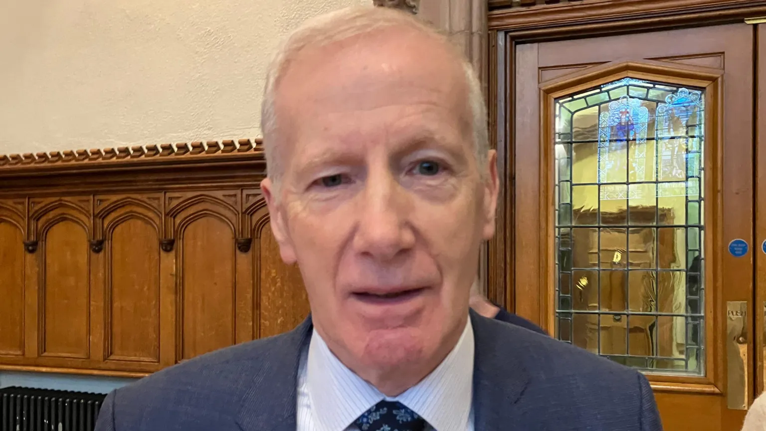 Gregory Campbell blue jacket, light-coloured shirt and dark floral tie in the main hall of the Guildhall. Wood panelling can be seen behind him as well as a wooden door with stained glass window.