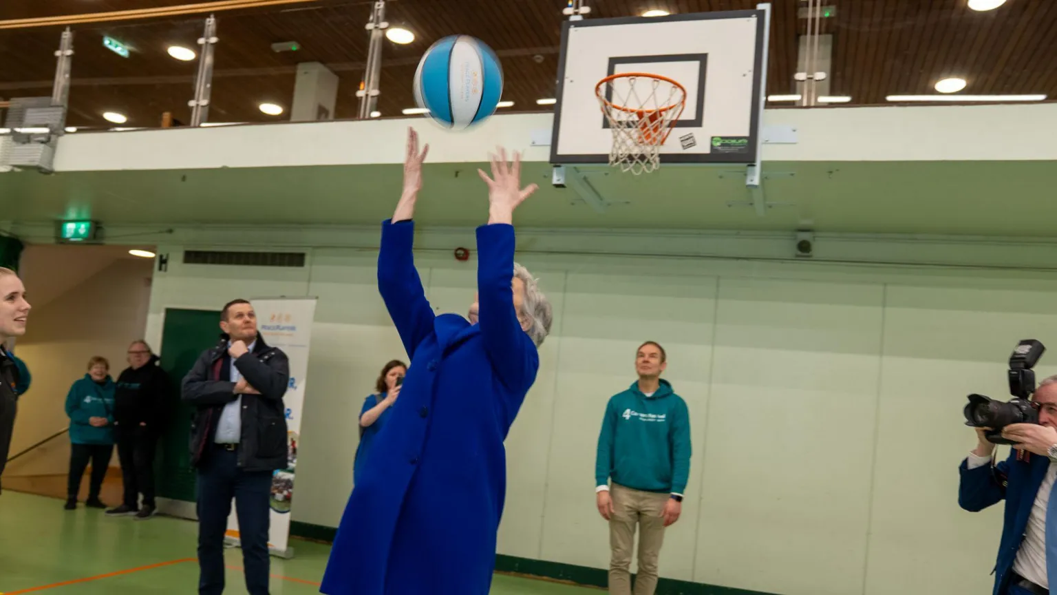 QUB Catherine Connolly in a blue jacket throwing a blue and white basketball over her head towards a basketball hoop. 