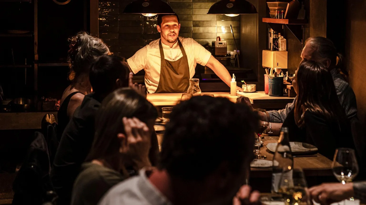 Eleonora Boscarelli Chef Corrin in kitchen which is open to dining area talking to diners at two tables together. The setting is dark with warm overhead lighting and the diners are all looking at him.