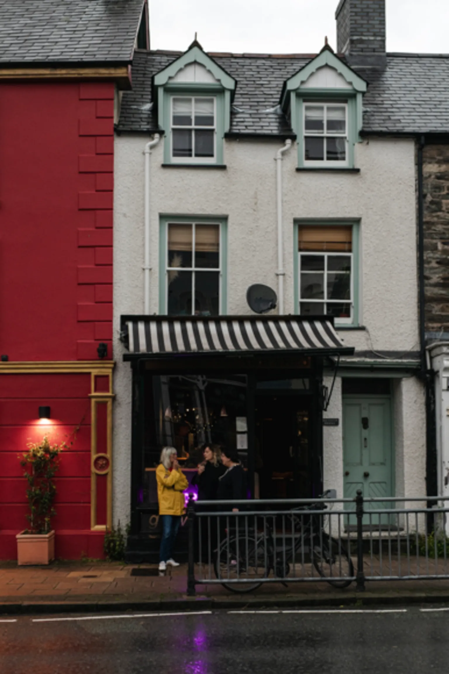 Eleonora Boscarelli Exterior of restaurant seen from across the road. The building is a narrow terraced house painted white with two dormer windows on the second floor, sash windows on the first floor and a shop front with striped awning on the ground floor next to a sage green front door. It is located next to a building which has been painted red.