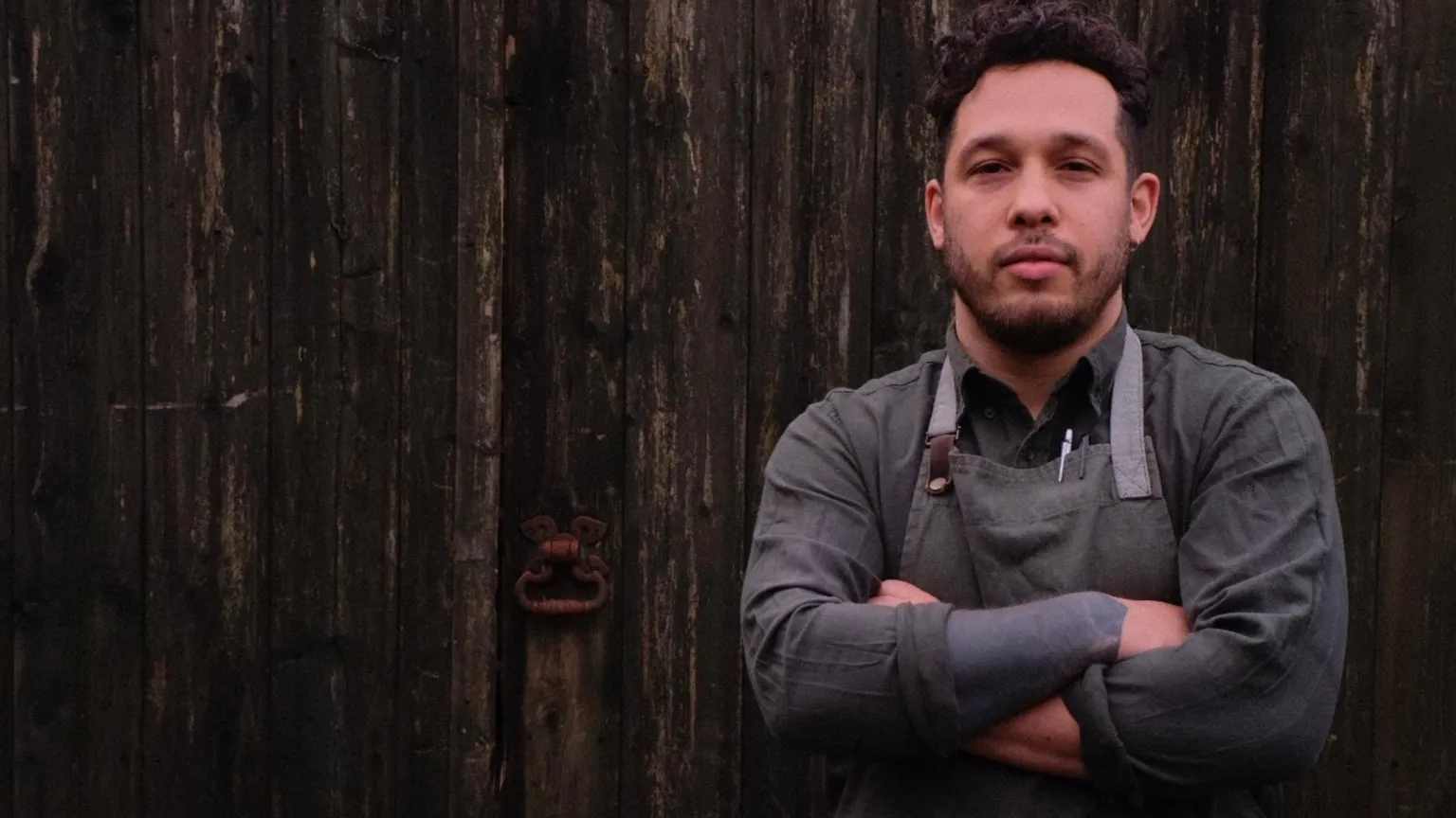 Abbie Morris Corrin Harrison in grey apron and dark grey shirt and arms crossed. He is standing in front of a wooden door and has dair hair and short trimmed dark facial hair