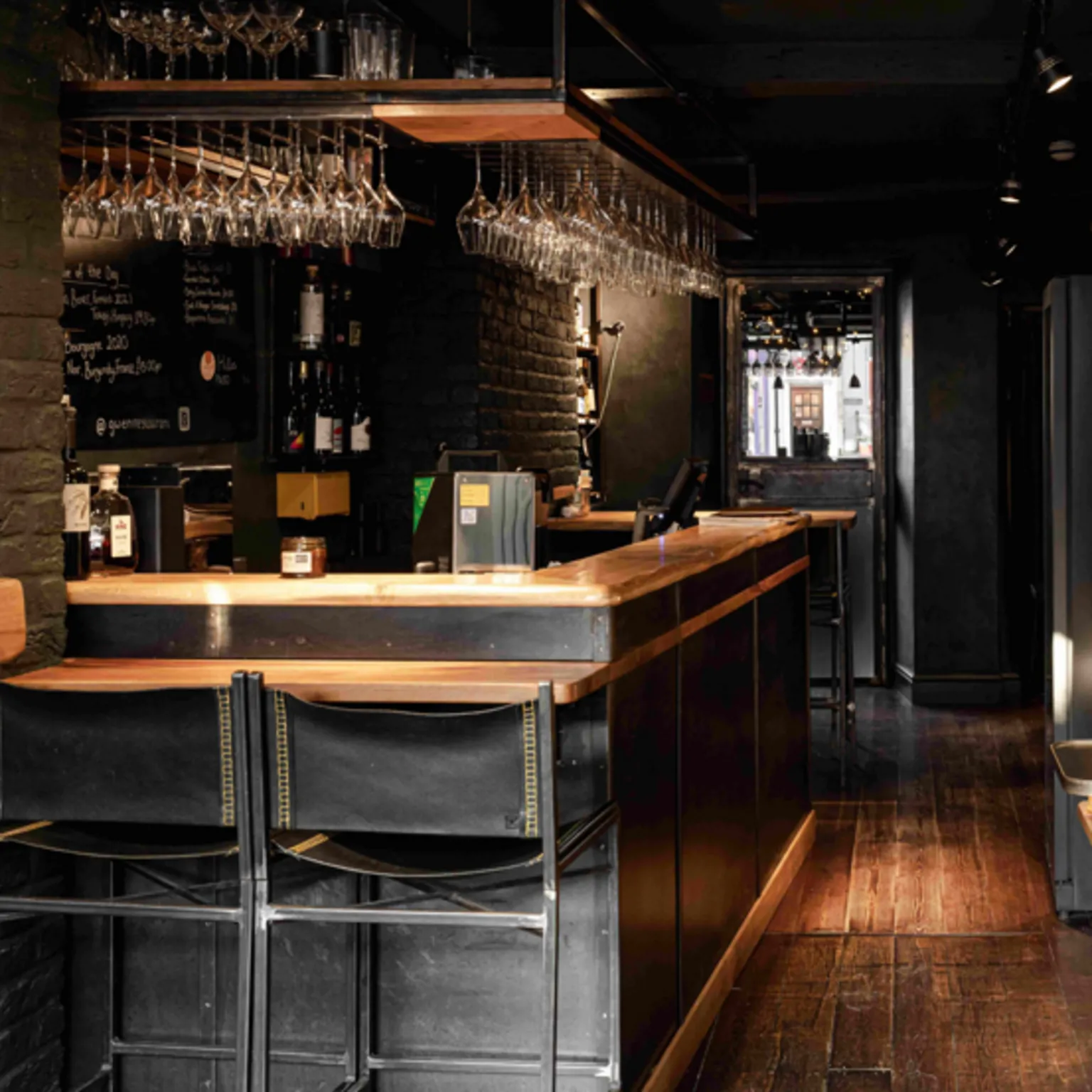 Emilia Boscarelli The bar area at the restaurant with dark walls and dark brown wooden flooring. Two black leather bar stools are tucked under the bar at the front and wine glasses hang from an overhead rack all around.