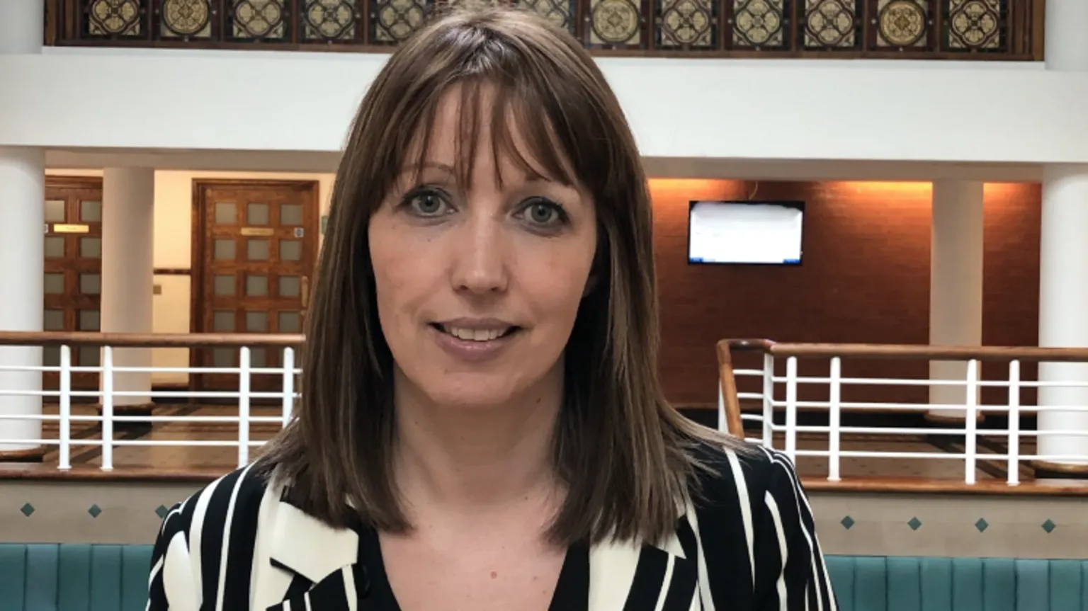 Sarah Sarah, with brown hair and fringe, wears a black-and-white shirt in an official-looking public building.